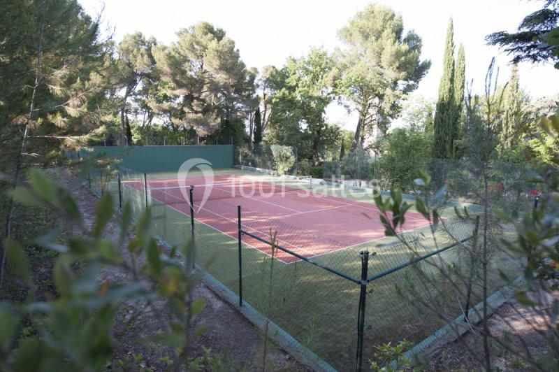 Terrain de tennis en plein air entouré de végétation et d'arbres, avec une clôture grillagée.