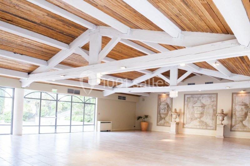Salle lumineuse avec poutres apparentes en bois, grandes baies vitrées et murs décorés de tableaux classiques.