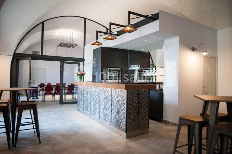 Salle moderne avec comptoir en métal décoratif, tabourets hauts, éclairage suspendu et vue sur une salle de réunion vitrée.