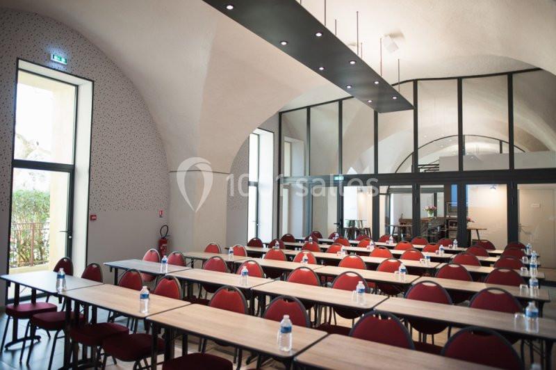 Salle de conférence lumineuse avec des tables alignées, des chaises rouges et une grande baie vitrée en arrière-plan.