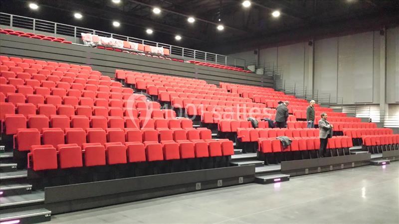 Gradins d'une salle de spectacle avec des sièges rouges et quelques personnes debout sur le côté.