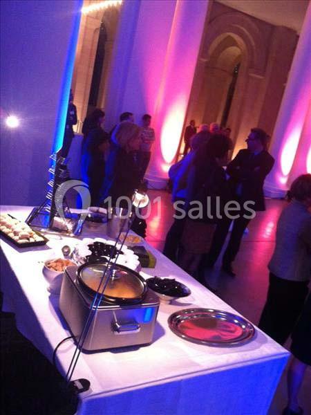 Table de buffet avec plats chauds et froids, dans une salle lumineuse avec colonnes et invités debout en arrière-plan.