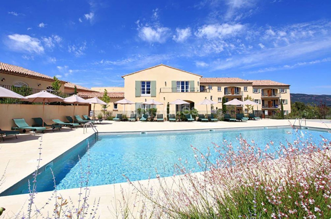 Piscine extérieure entourée de chaises longues et parasols devant un bâtiment beige sous un ciel bleu dégagé.
