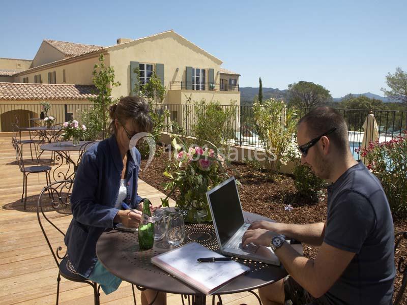 Deux personnes travaillent sur une terrasse ensoleillée, entourées de plantes et de bâtiments provençaux.