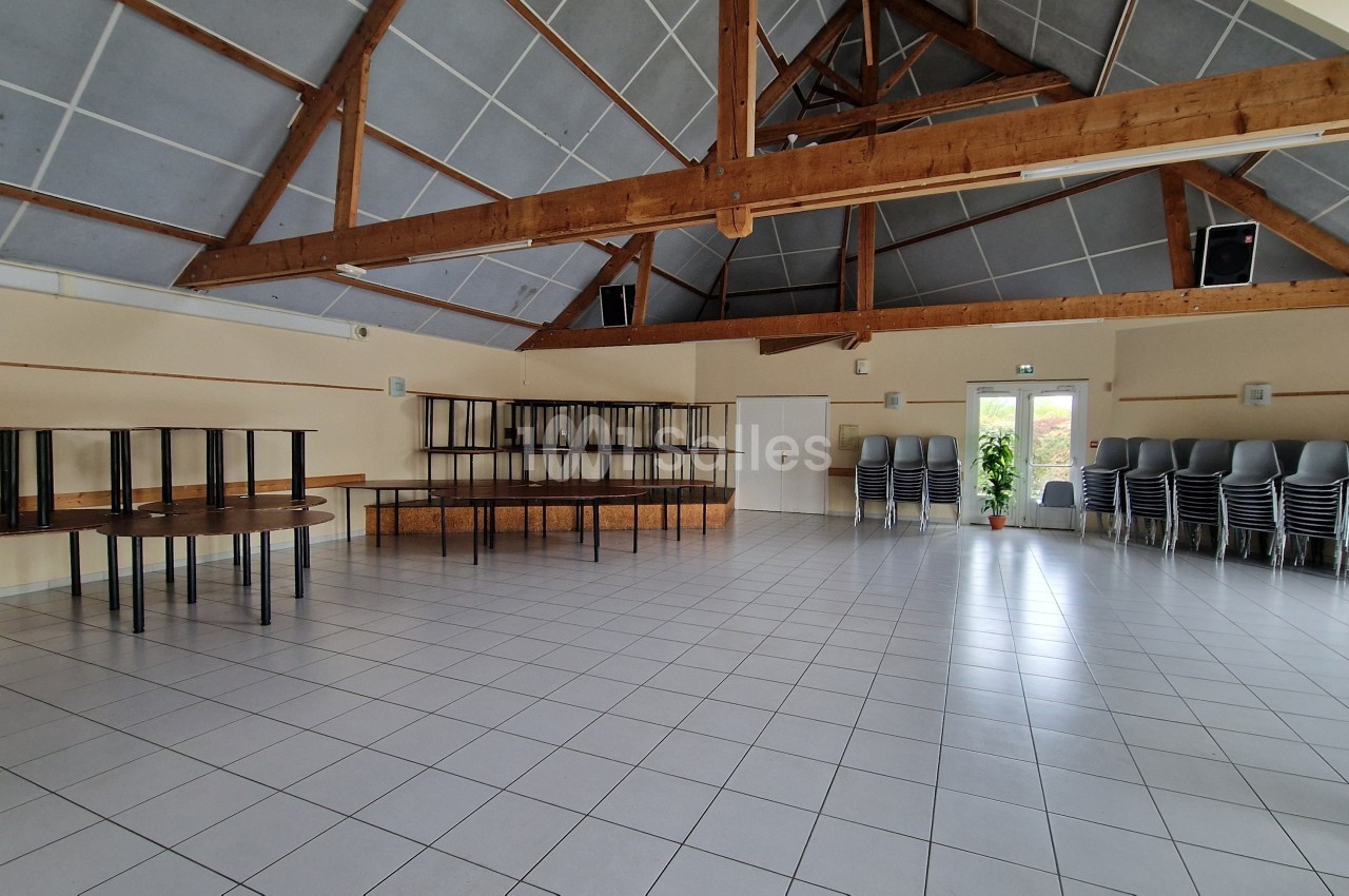 Salle vide avec un sol carrelé, des poutres apparentes, des tables empilées et des chaises rangées contre un mur.