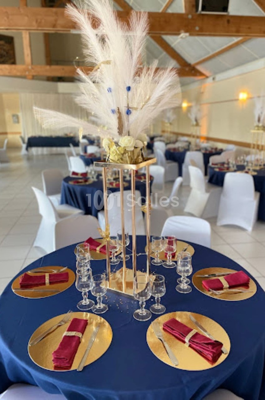 Salle de réception décorée avec des tables rondes, nappes bleues, serviettes rouges et centre de table floral élégant.