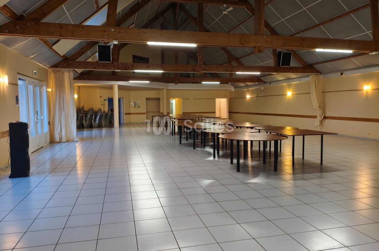Salle spacieuse avec charpente en bois apparente, tables alignées et chaises empilées dans un coin.
