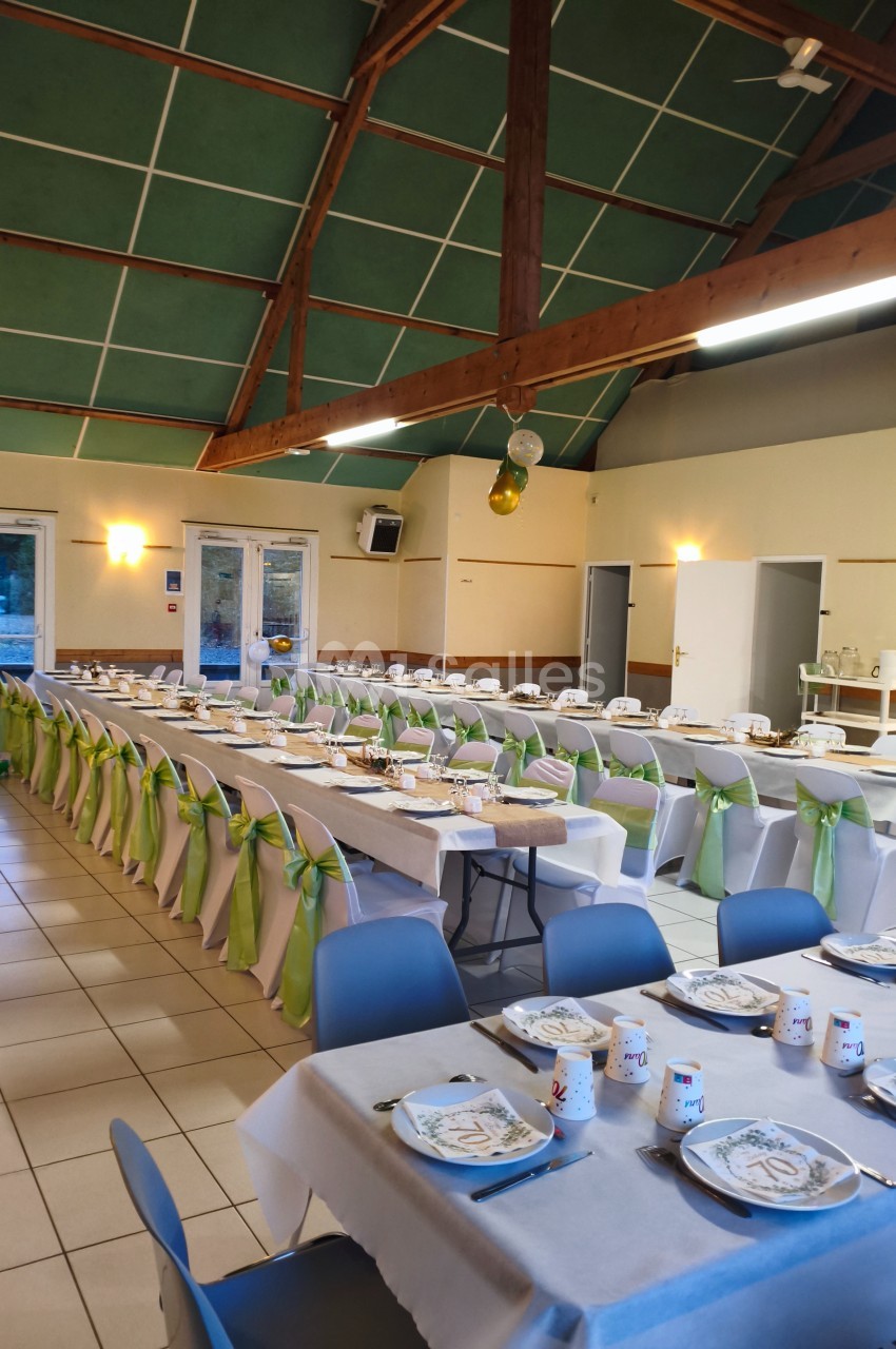 Salle de réception décorée avec des tables alignées, nappes blanches, chaises ornées de rubans verts.