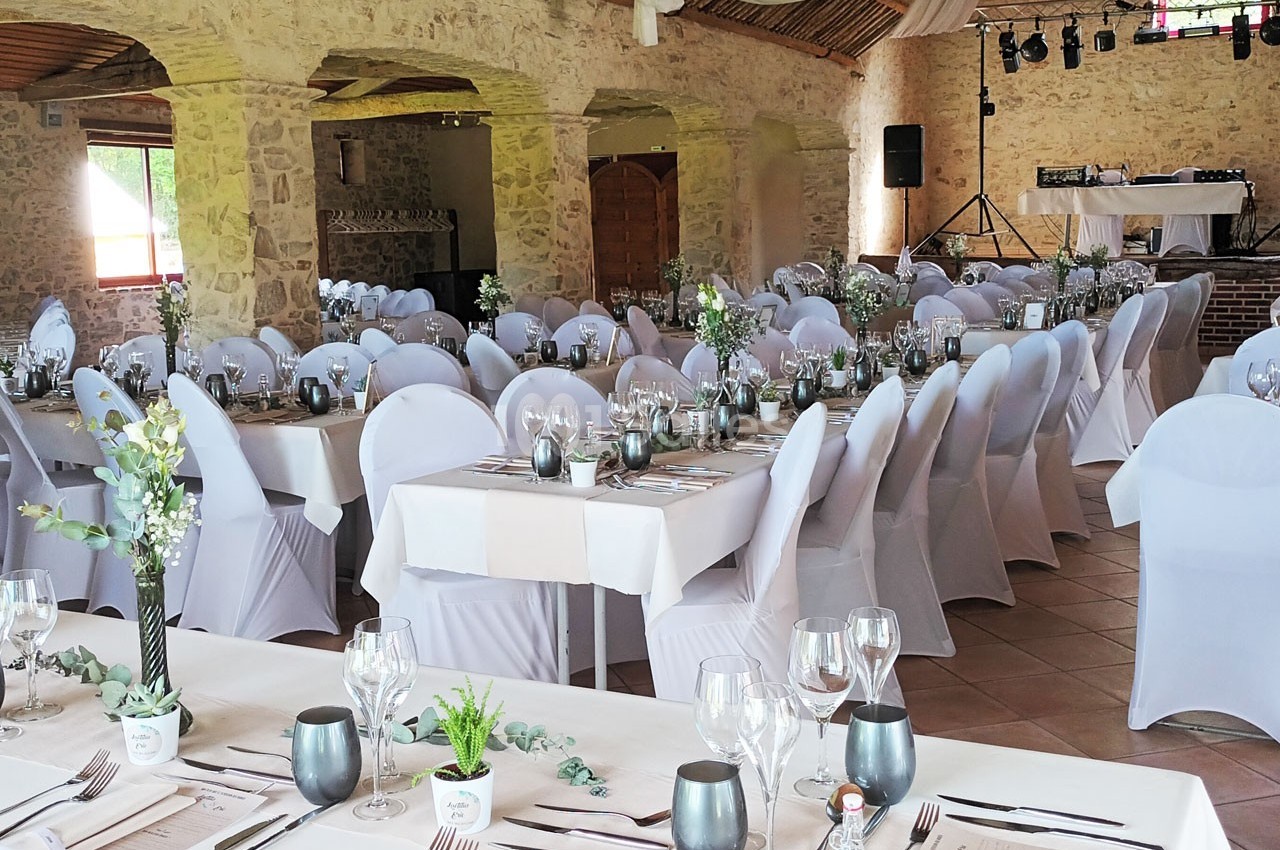 Salle de réception décorée pour un événement, avec des tables dressées, des chaises blanches et des arrangements floraux.