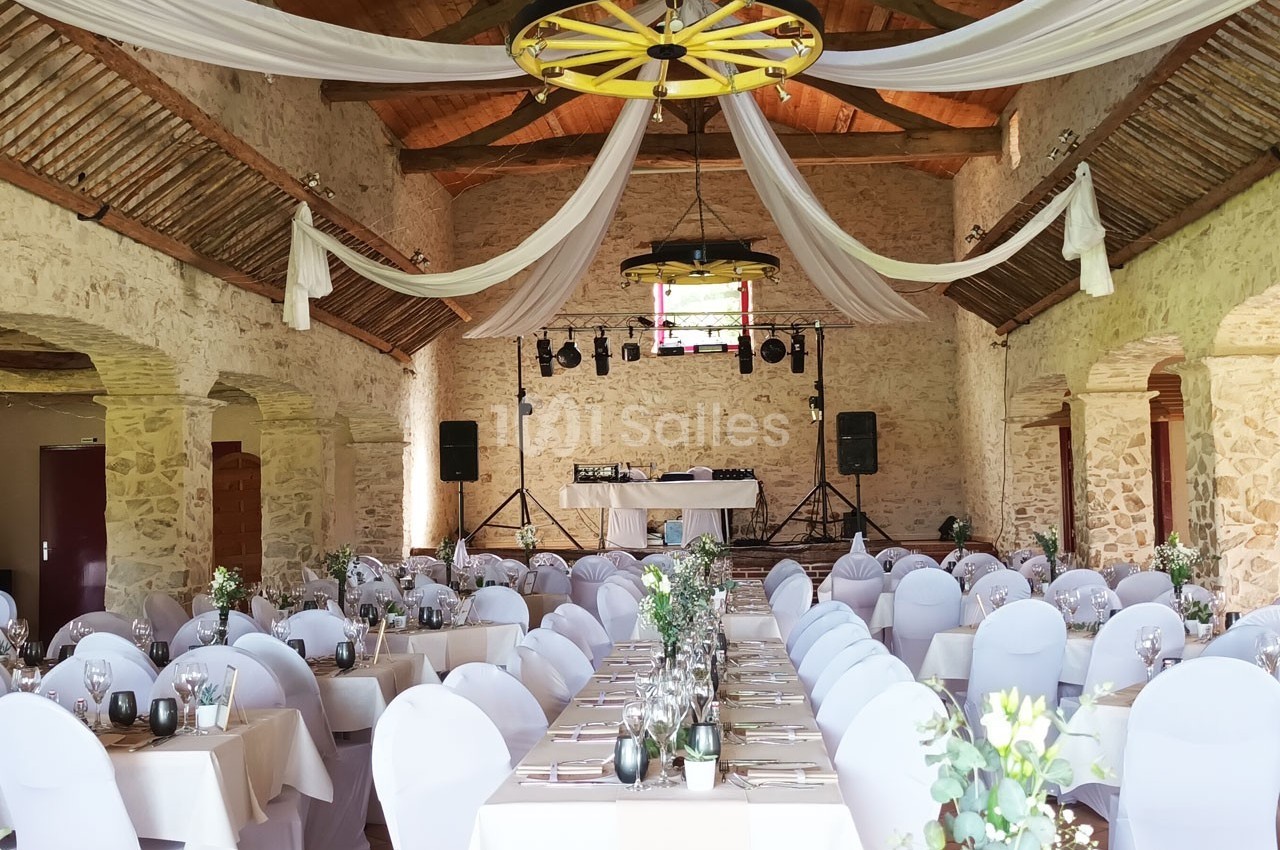 Salle de réception en pierre avec tables décorées, chaises blanches et drapés suspendus au plafond.