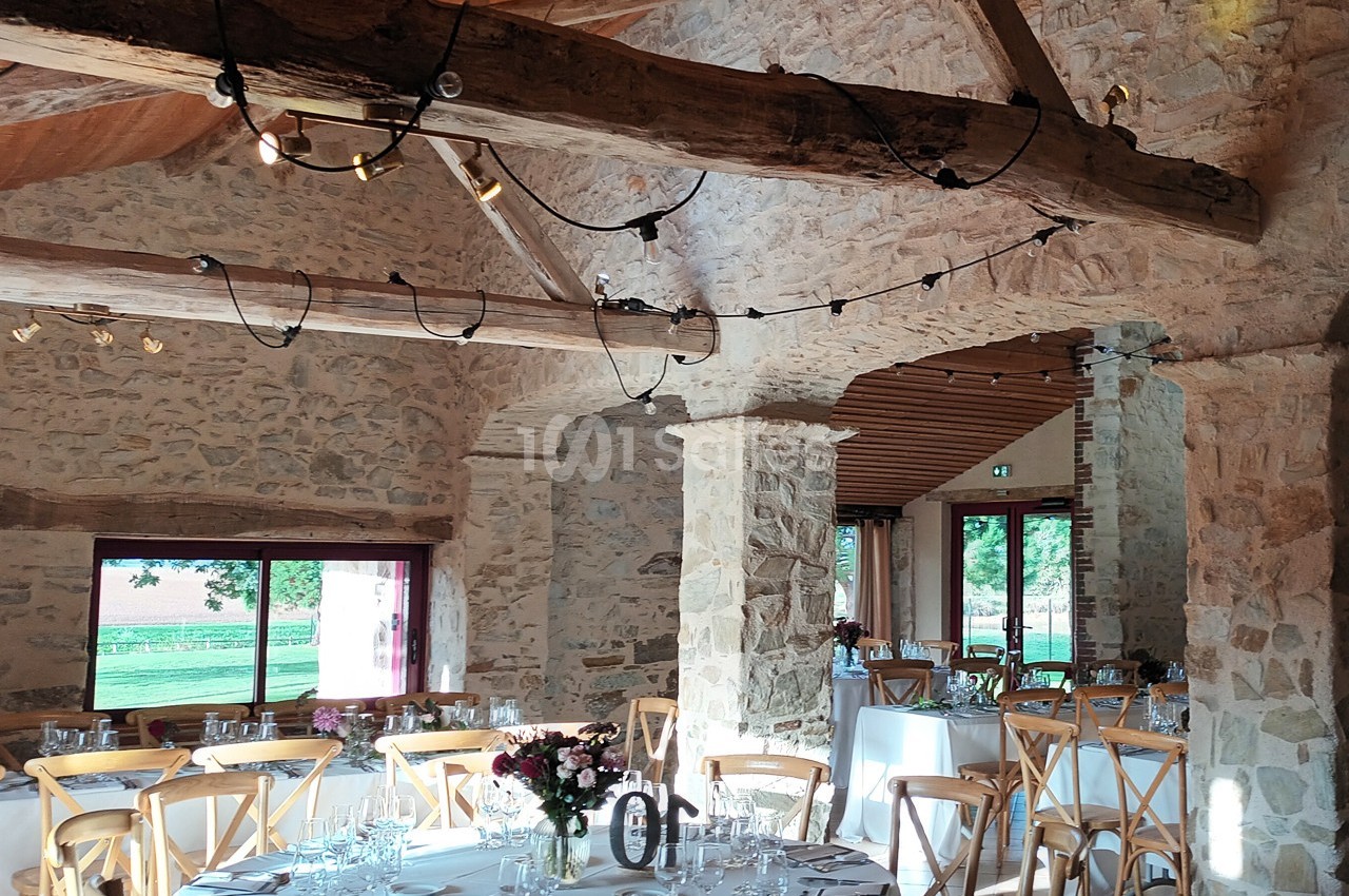 Salle en pierre avec poutres apparentes, tables dressées pour un repas, lumière naturelle entrant par les fenêtres.