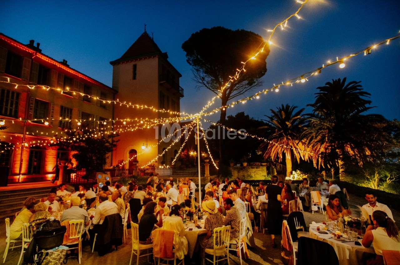 Dîner en plein air sous des guirlandes lumineuses, avec des invités assis autour de tables dans un cadre festif.