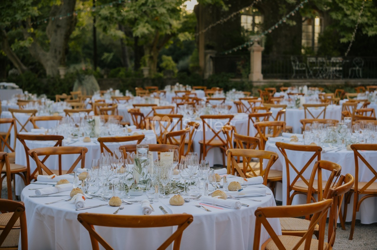 Tables rondes dressées avec nappes blanches, chaises en bois, vaisselle et décoration florale dans un jardin extérieur.