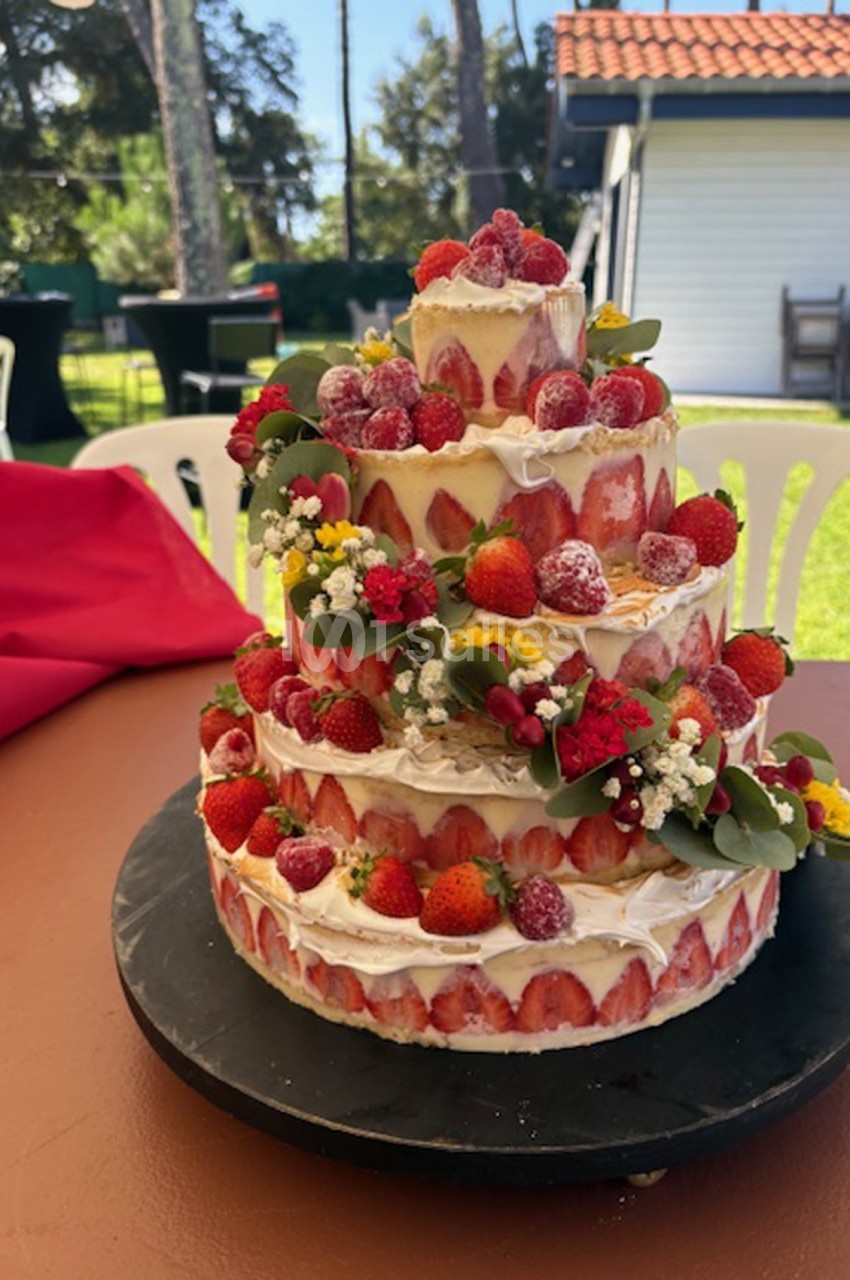 Gâteau à étages décoré de fraises, framboises et fleurs, présenté sur une table en extérieur par temps ensoleillé.