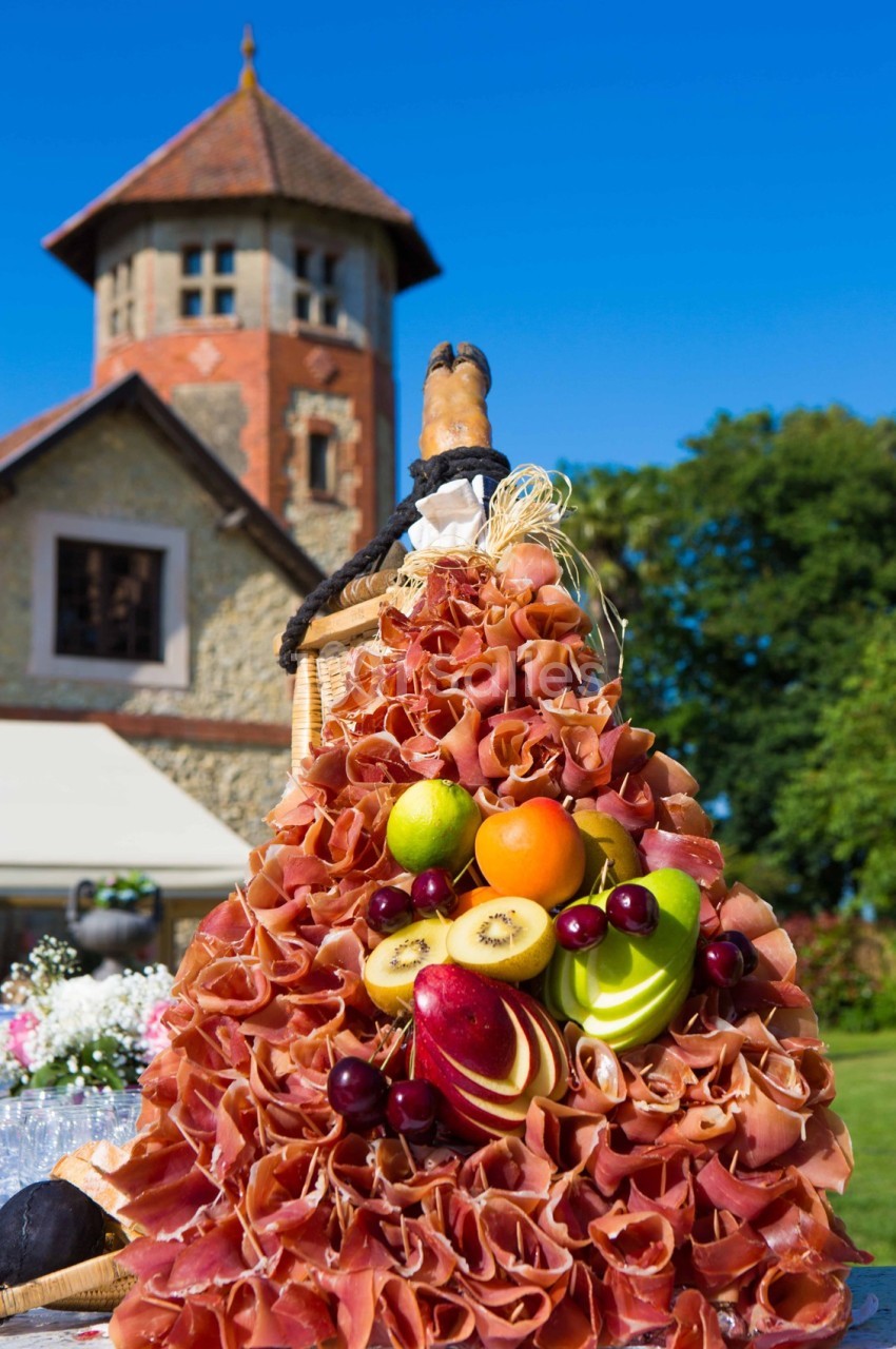 Plateau de charcuterie en forme de pyramide décoré de fruits frais, avec une maison en pierre et un ciel bleu en arrière…
