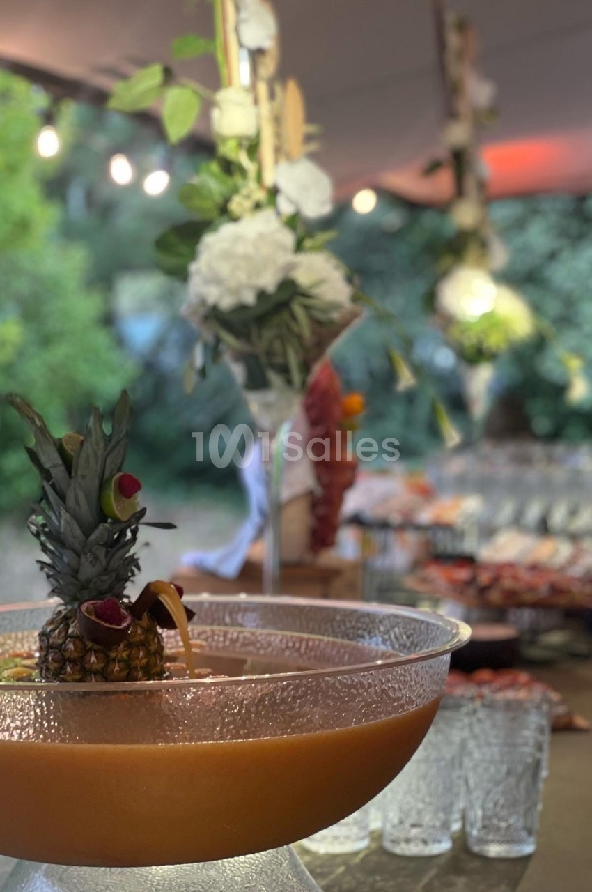 Grand bol de cocktail tropical décoré d’un ananas, sur une table dressée en extérieur avec des lumières et fleurs.