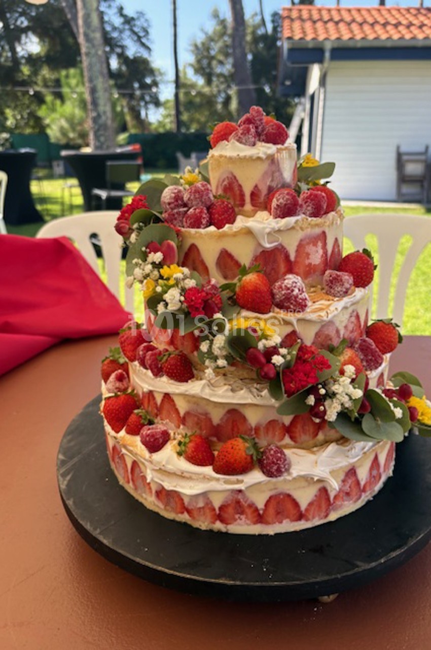 Gâteau à étages décoré de fraises, framboises et fleurs, présenté sur une table en extérieur par temps ensoleillé.