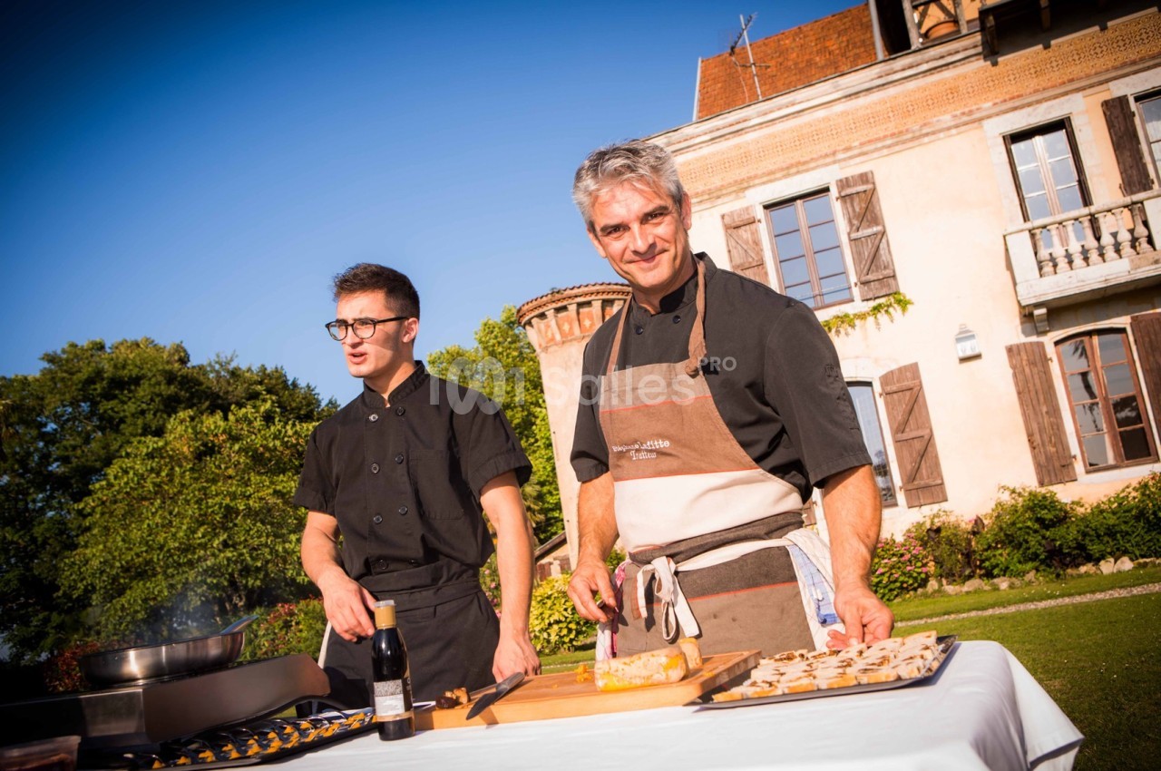 Deux chefs cuisinent en extérieur devant une grande maison, avec des ingrédients disposés sur une table.