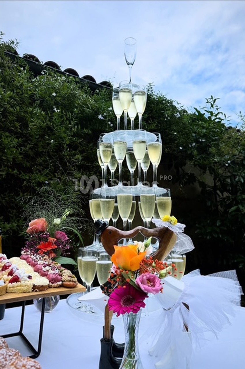 Tour de coupes de champagne empilées en pyramide sur une table décorée de fleurs et de desserts, en extérieur.