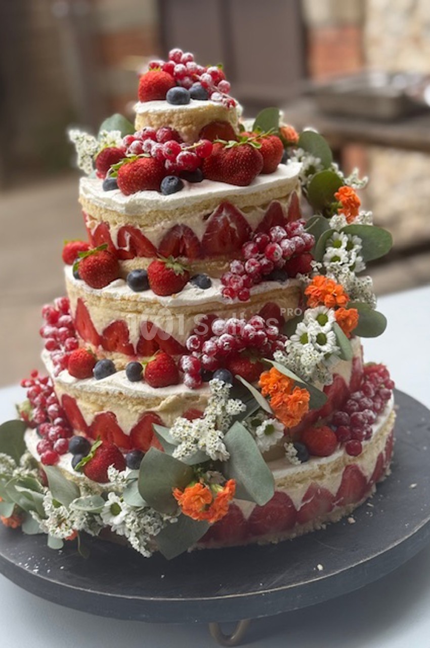 Gâteau de mariage à étages décoré de fruits rouges, baies, fleurs fraîches et feuillage, présenté sur un plateau noir.