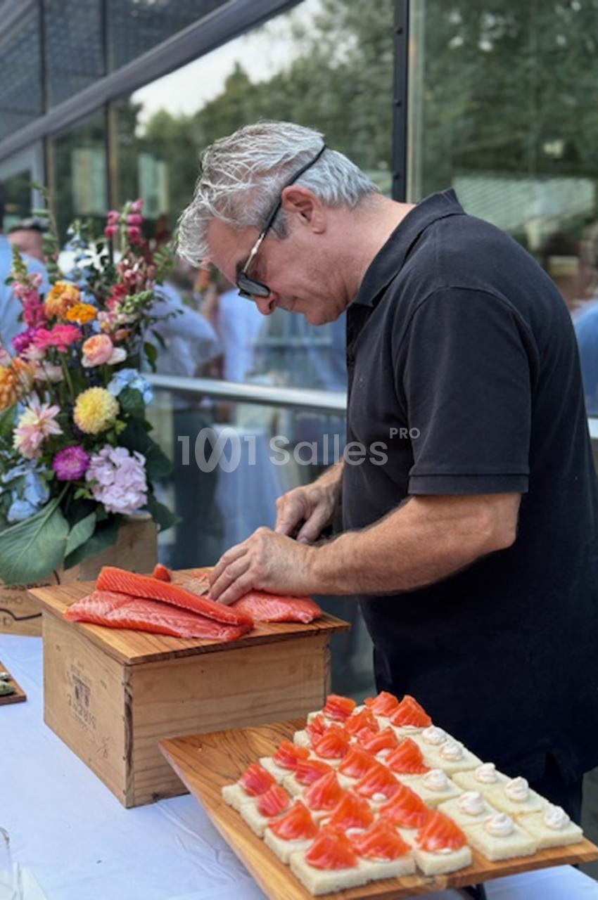 Un homme découpe du saumon frais sur une planche en bois, à côté de fleurs et de bouchées apéritives.