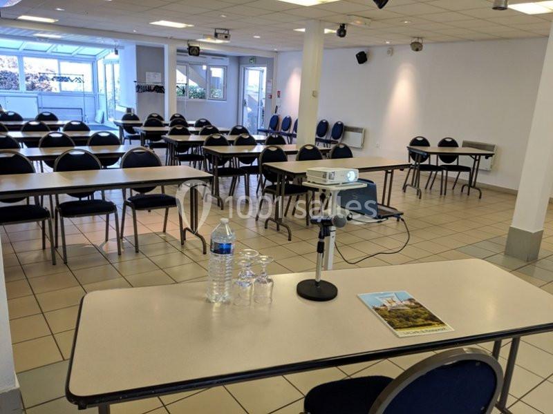 Salle de conférence lumineuse avec des tables, des chaises alignées et un projecteur installé sur une table à l'avant.