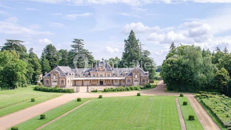 Manoir entouré de jardins verdoyants, avec allées gravillonnées et arbres en arrière-plan sous un ciel partiellement nuageux.