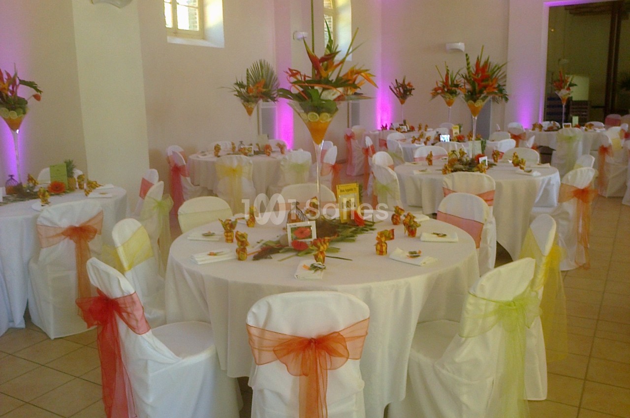 Salle décorée pour un événement avec des tables rondes, nappes blanches, chaises ornées de rubans colorés et centres de…
