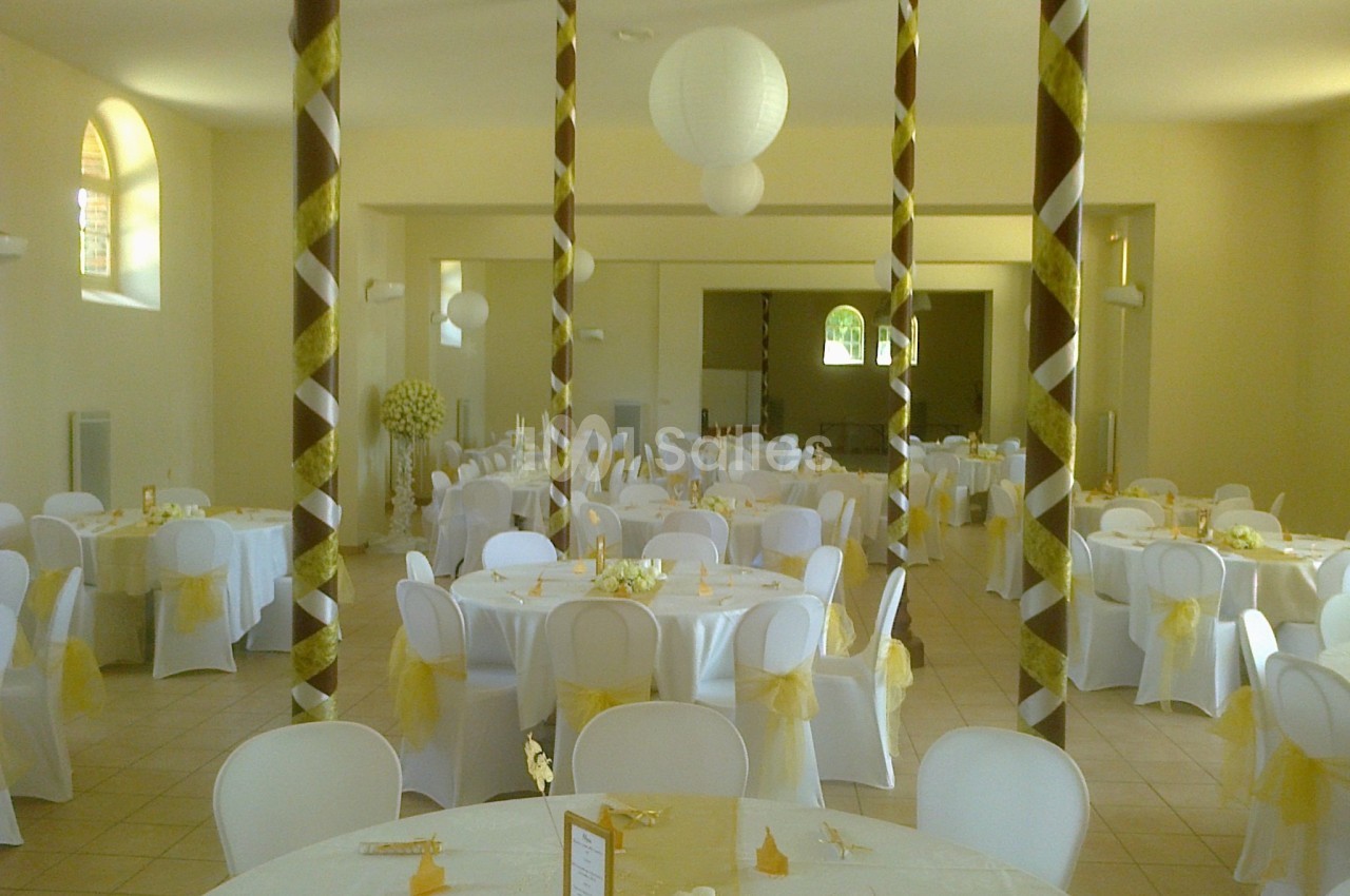 Salle de réception décorée avec des tables rondes, nappes blanches, chaises assorties et ornements dorés.