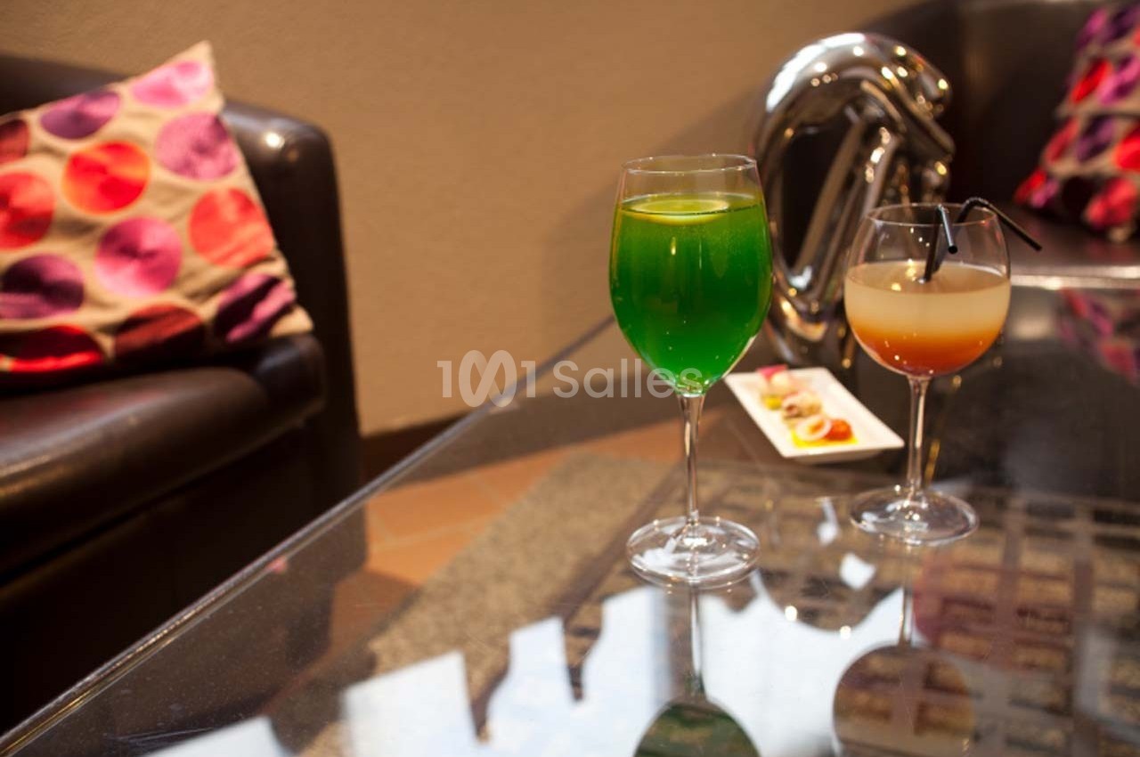 Deux verres de cocktails colorés posés sur une table en verre, avec un canapé et des coussins en arrière-plan.