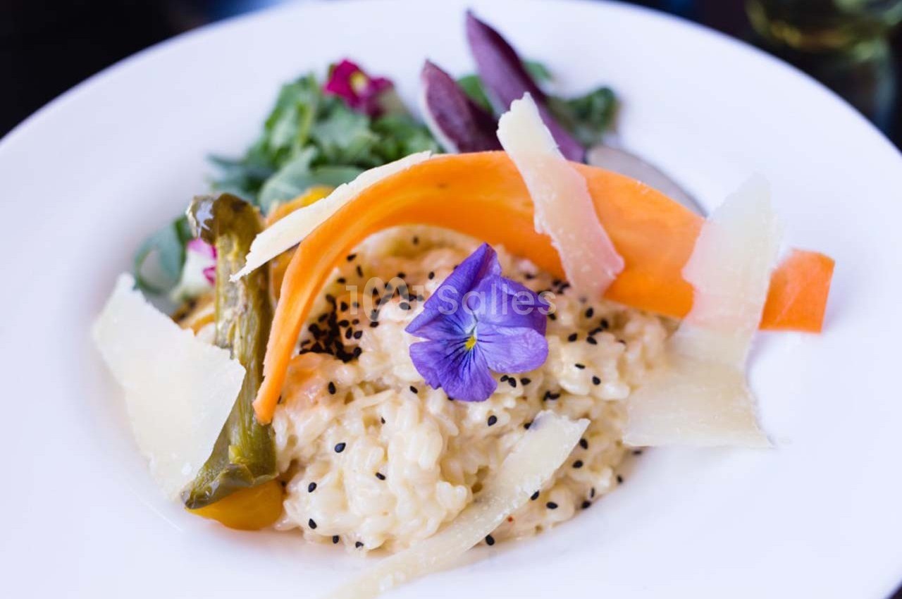 Assiette de risotto crémeux garni de légumes colorés, copeaux de parmesan et une fleur comestible violette.