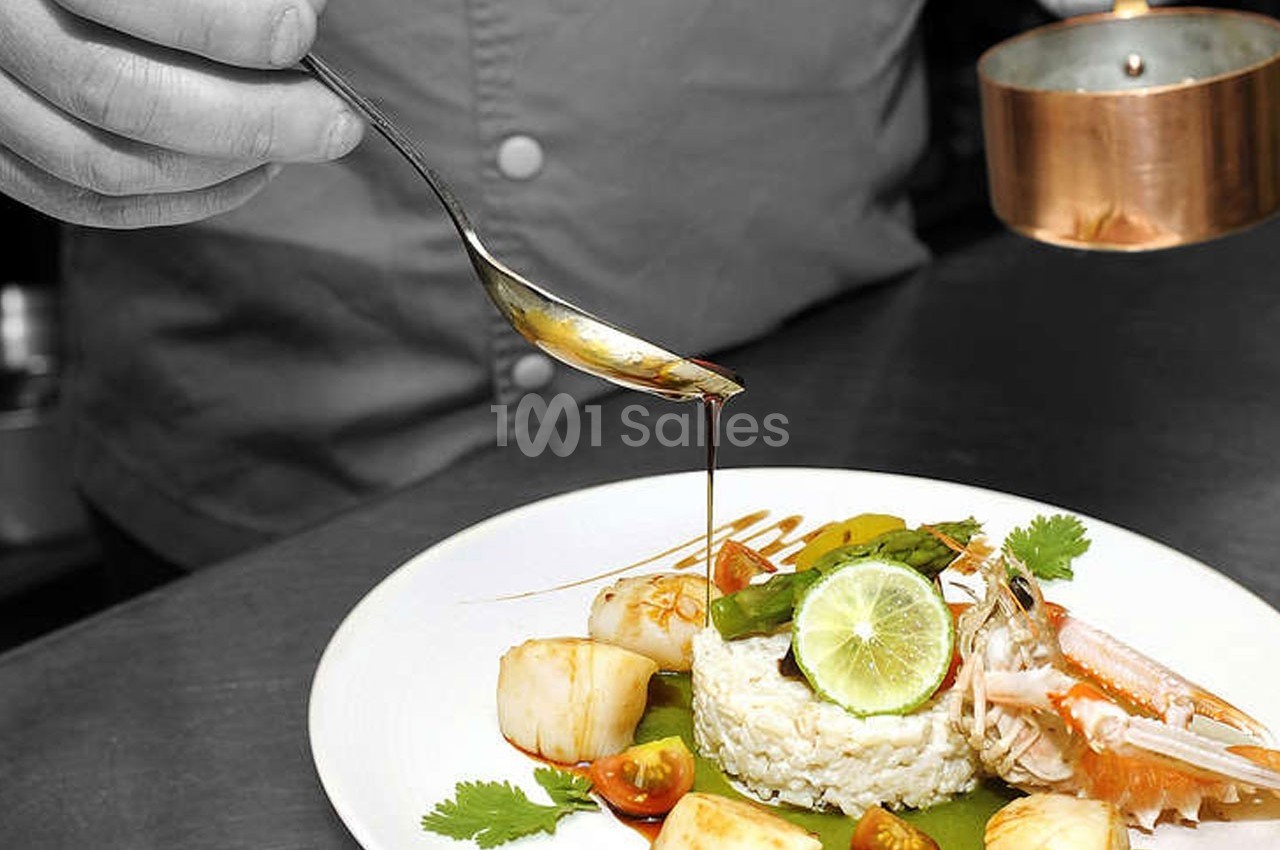 Un chef verse une sauce sur un plat composé de riz, noix de Saint-Jacques, crevette, légumes et citron vert.