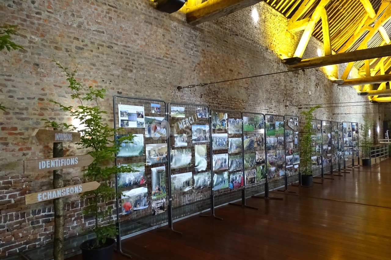 Exposition de photos accrochées sur des grilles dans une grande salle en briques avec un plafond en poutres apparentes.