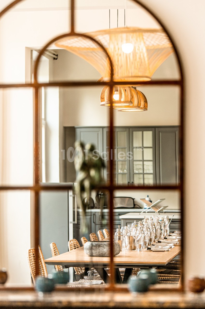Vue d'une salle à manger élégante avec une table dressée, des chaises en osier et une cuisine en arrière-plan.