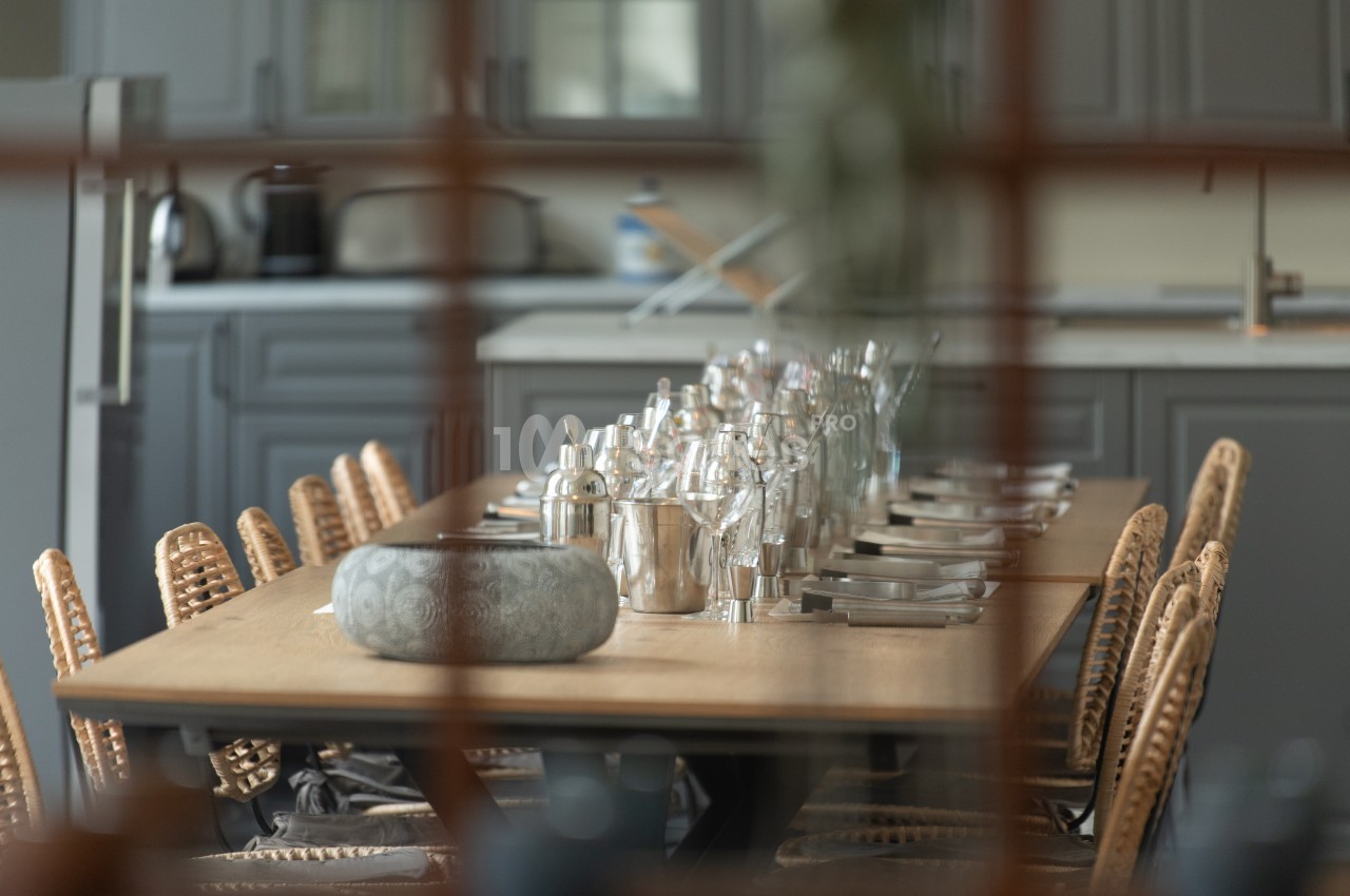 Table en bois dressée avec des couverts, des verres et des carafes, entourée de chaises en rotin dans une cuisine moderne.