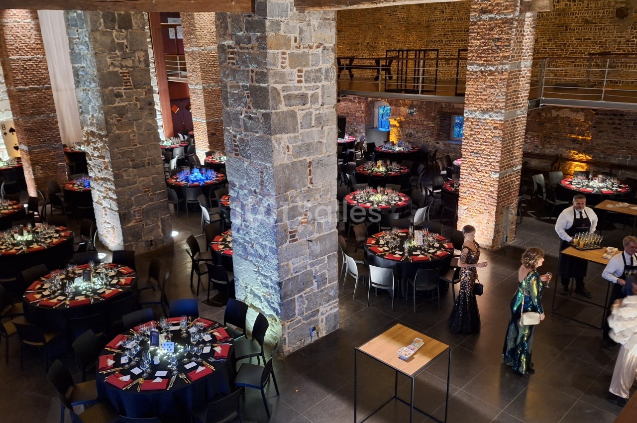 Salle en pierre avec tables rondes dressées pour un événement, éclairage tamisé et quelques personnes en tenue de soirée.