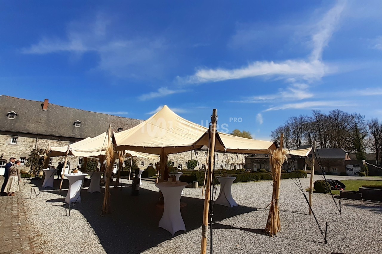 Tente beige installée dans une cour pavée, entourée de tables hautes et de bâtiments en pierre sous un ciel bleu.