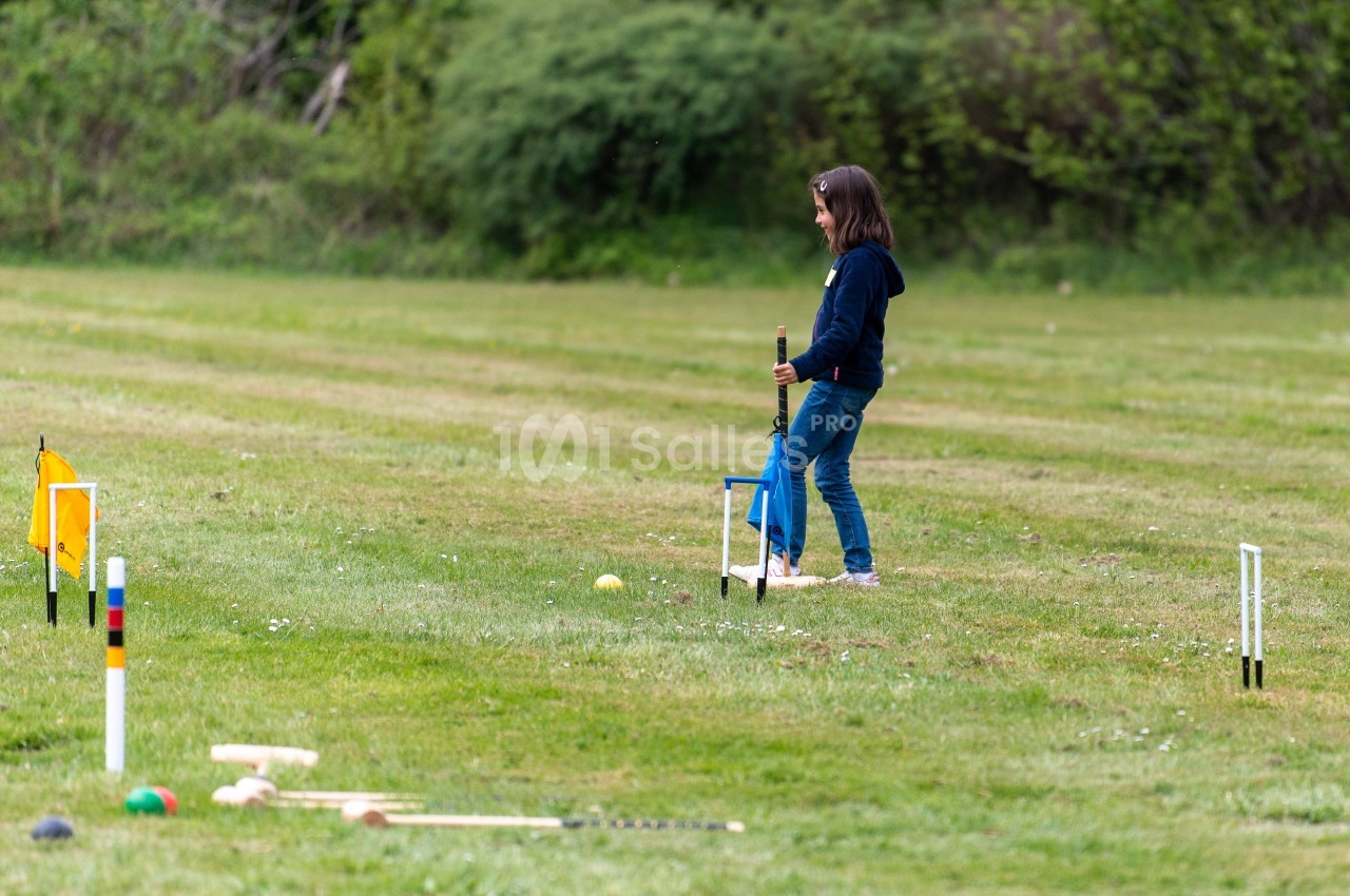Une jeune fille joue au croquet sur une pelouse, entourée de piquets et de maillets.