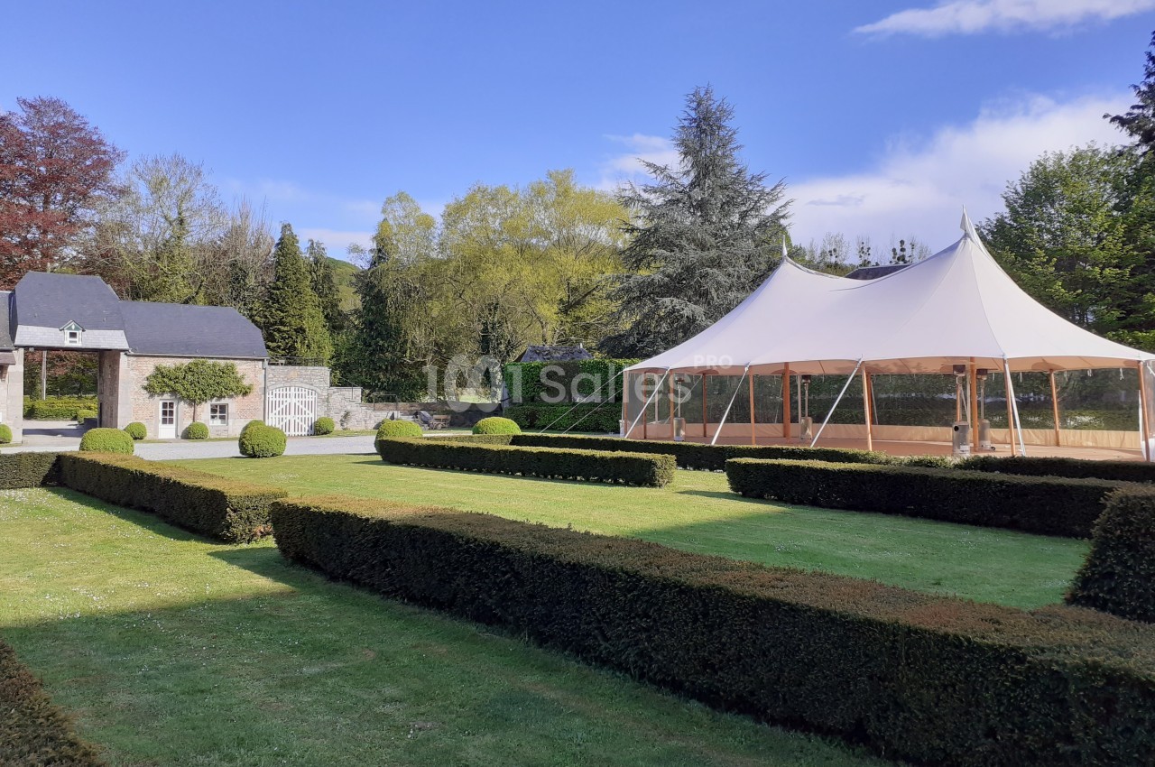 Jardin avec pelouse, haies taillées, tente blanche et bâtiment en pierre sous un ciel bleu.