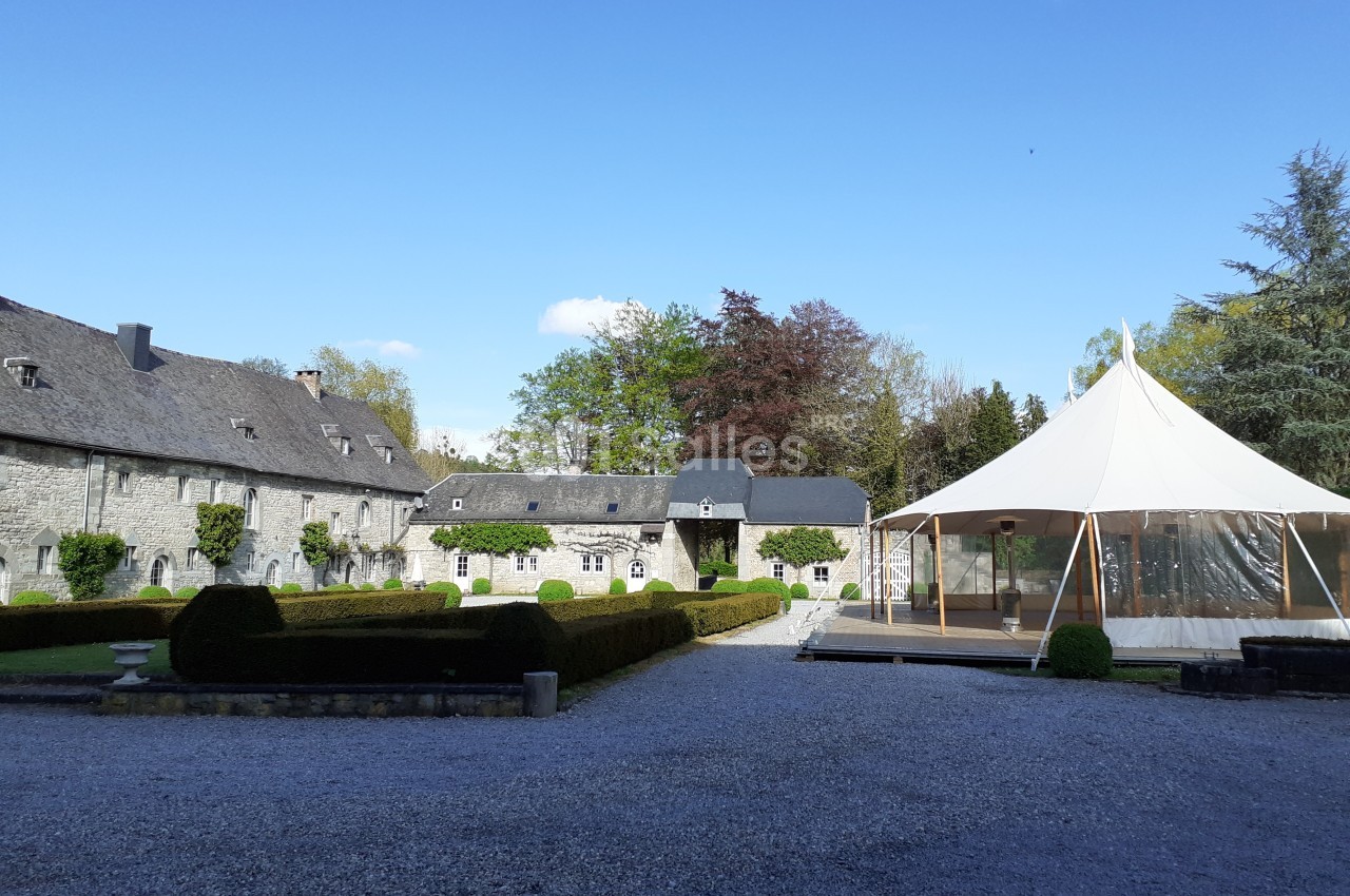 Cour d’un domaine avec bâtiments en pierre, haies taillées et une tente blanche sur une terrasse en bois.