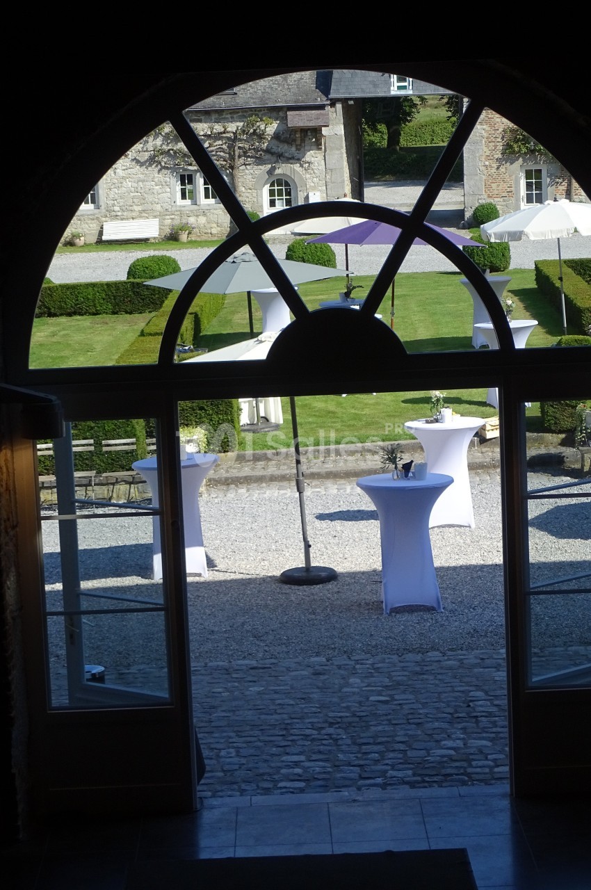 Vue depuis une porte vitrée sur une cour pavée avec des tables hautes dressées et un jardin en arrière-plan.