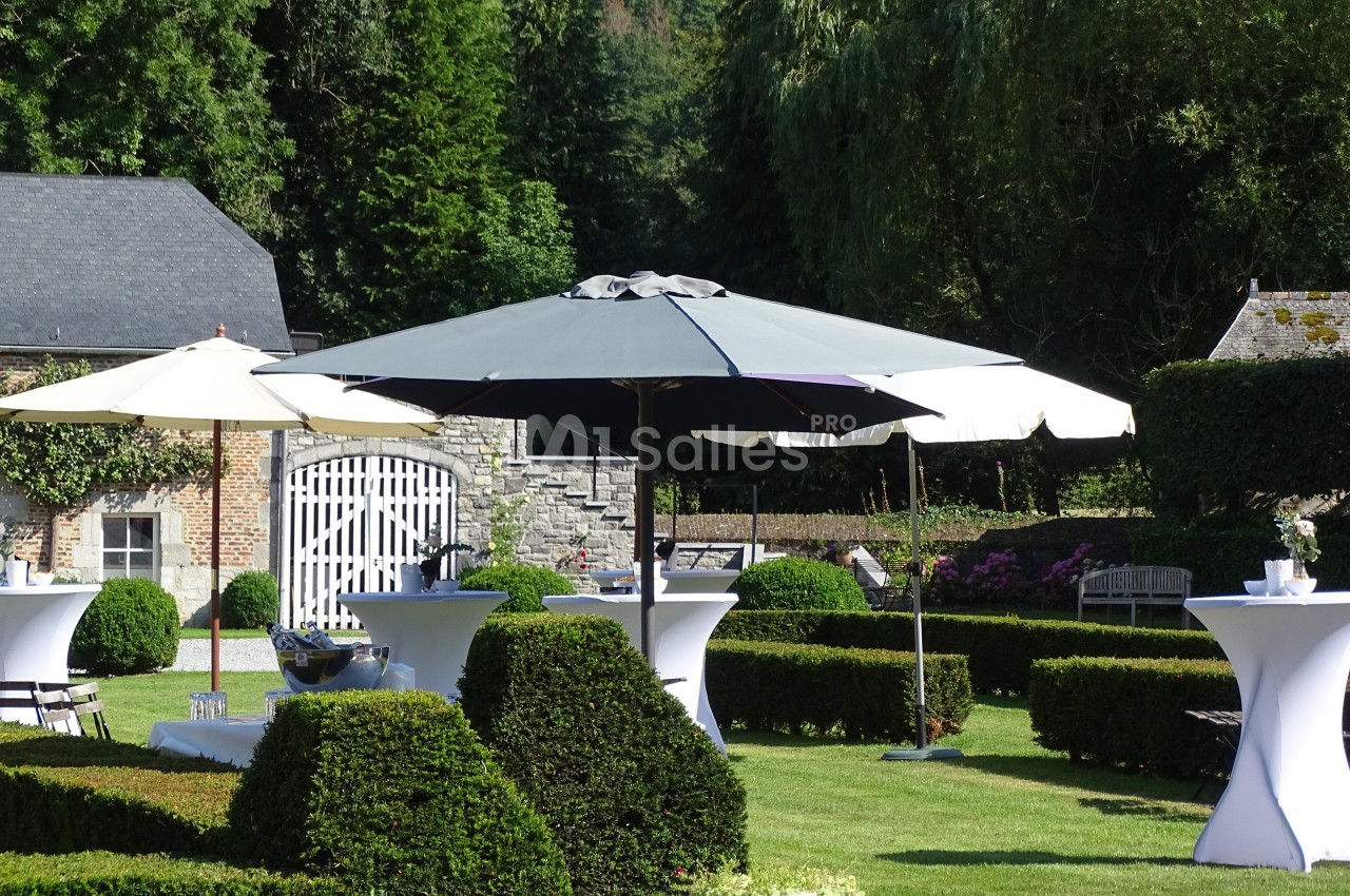Parasols blancs et gris installés dans un jardin verdoyant avec des tables hautes et des haies taillées.