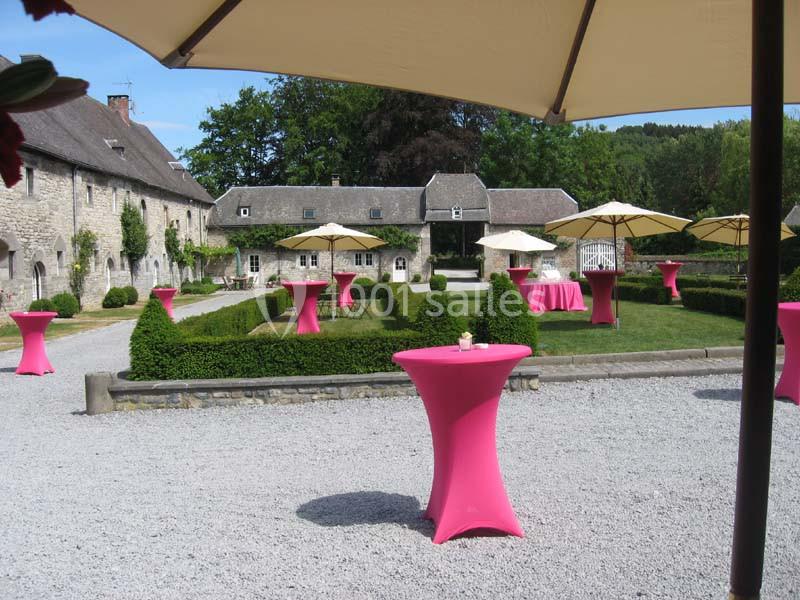 Cour extérieure d'un domaine avec des tables hautes roses et des parasols beige, entourée de verdure et bâtiments en pierre.