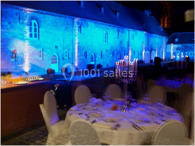 Salle de réception éclairée en bleu avec une table dressée au premier plan et un bâtiment en pierre en arrière-plan.