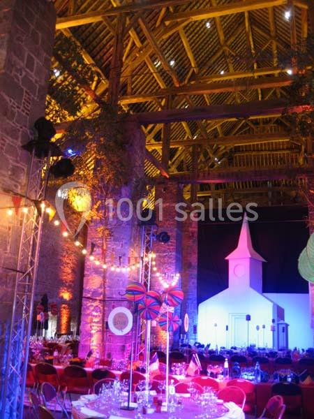 Intérieur d'une grange aménagée avec éclairages colorés, tables dressées et décorations festives, incluant une maquette d…