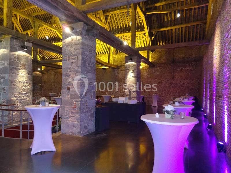 Salle en pierre avec poutres apparentes, tables hautes blanches et éclairage violet créant une ambiance chaleureuse.