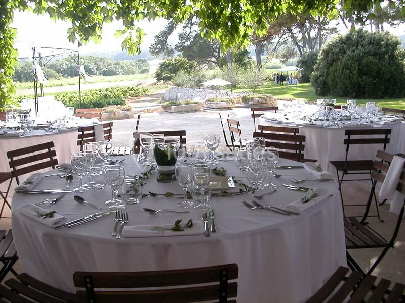 Tables rondes dressées avec nappes blanches, couverts et verres, disposées en extérieur sous des arbres.