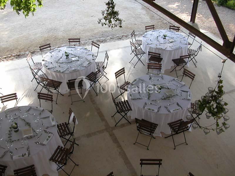 Tables rondes dressées avec nappes blanches et chaises pliantes en bois, disposées dans un espace extérieur couvert.