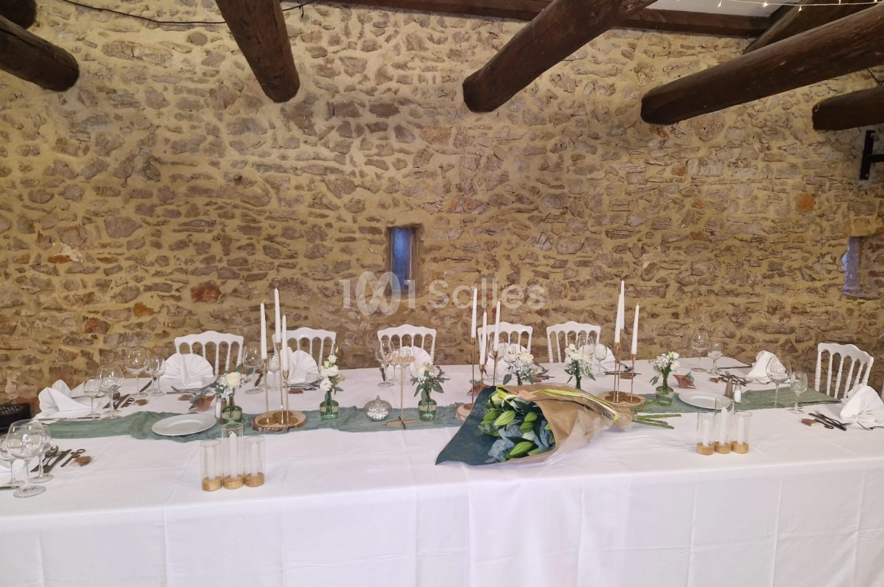 Table de réception décorée avec nappes blanches, bougies, vaisselle élégante et bouquet de fleurs, dans une salle en pierre.