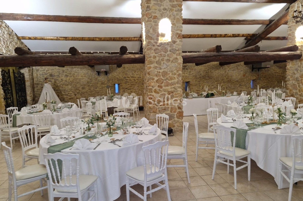 Salle de réception en pierre avec tables rondes dressées, chaises blanches et décoration élégante.